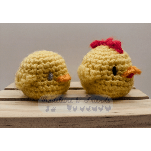 Two small, round crochet chickens, sitting next to each other, and facing right. They have no legs. On the left is a chick, with no wattle or comb, and blue eyes. On the right is an adult chicken. It is slightly larger, with a red wattle and comb, and black eyes. There is a see through watermark at the bottom. It says "Madeleine & Friends" and shows blue silhouettes of a line of rabbits and chickens wearing various capes and hats, or carrying fantasy and scifi weapons. 