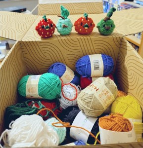 A box fall of 50g balls of cotton yarn from Ricorumi and Sirdar. On the open lid of the box are four little crochet chickens. Two look like little apples, with a stalk and leaf instead of a comb, and two look like strawberries.